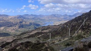 A mountainous landscape with a clear sky, a lake, and a trail with wooden posts.