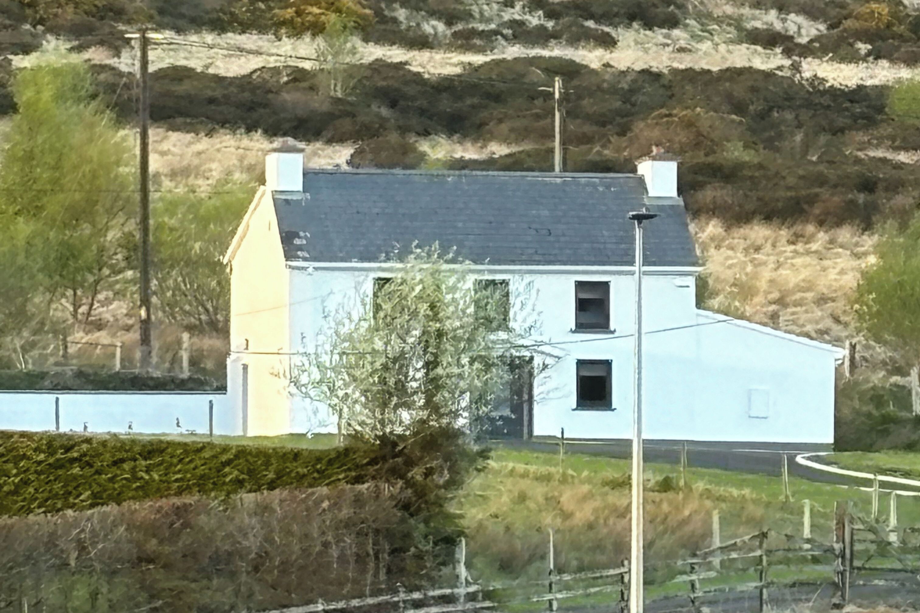 A white house with a grey roof, surrounded by a fenced garden and a hill in the background.