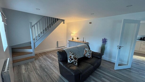 A modern living room with a black leather sofa, a wooden staircase, and a kitchen area visible through an open door.