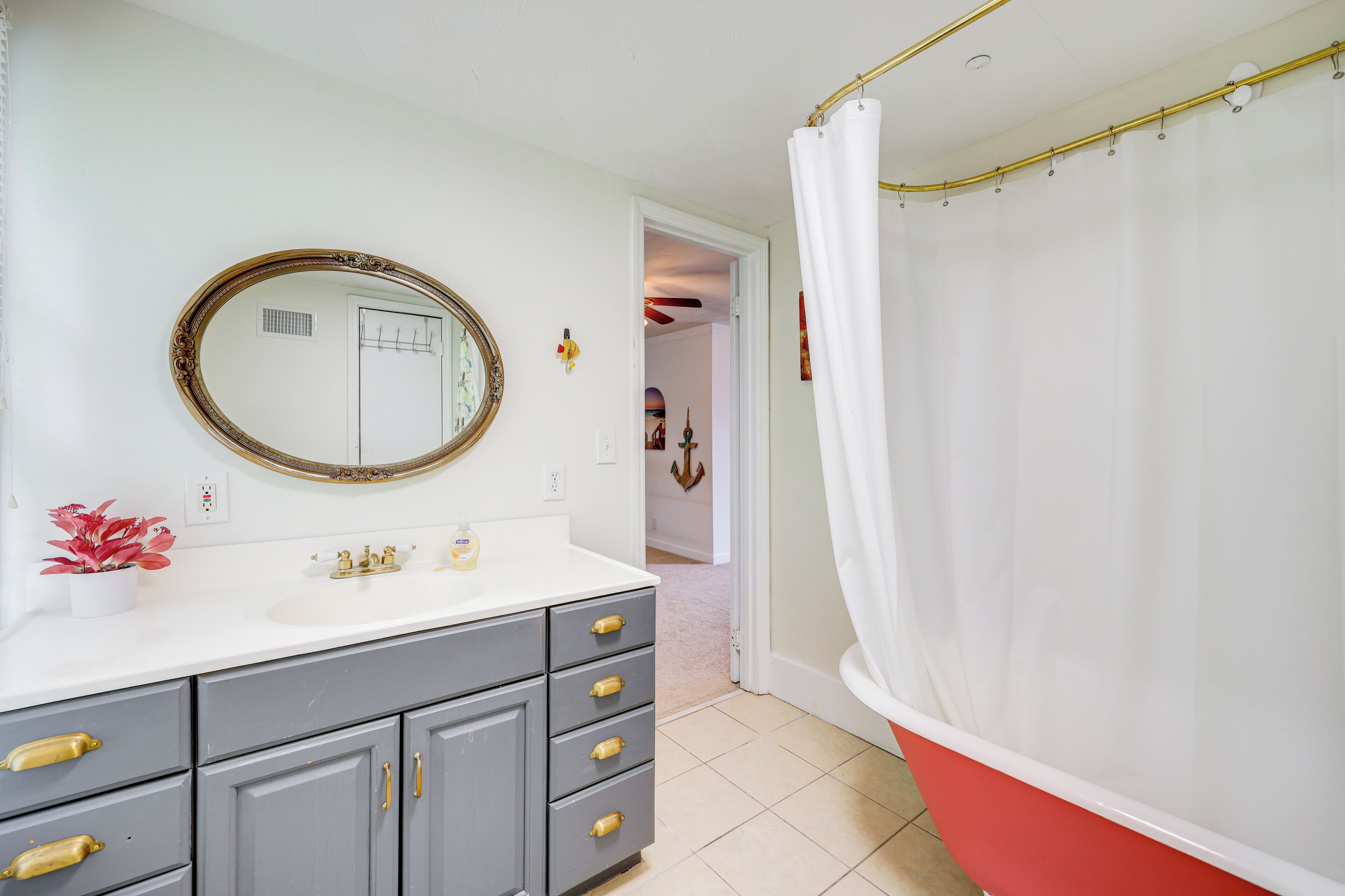 Un bagno con un lavandino bianco, uno specchio con una cornice decorata, mobili grigi con maniglie in ottone e una vasca da bagno rossa con tenda da doccia bianca.