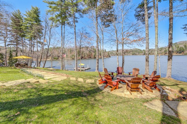 Un patio junto al lago con sillas Adirondack y una mesa, rodeado de árboles y un muelle.