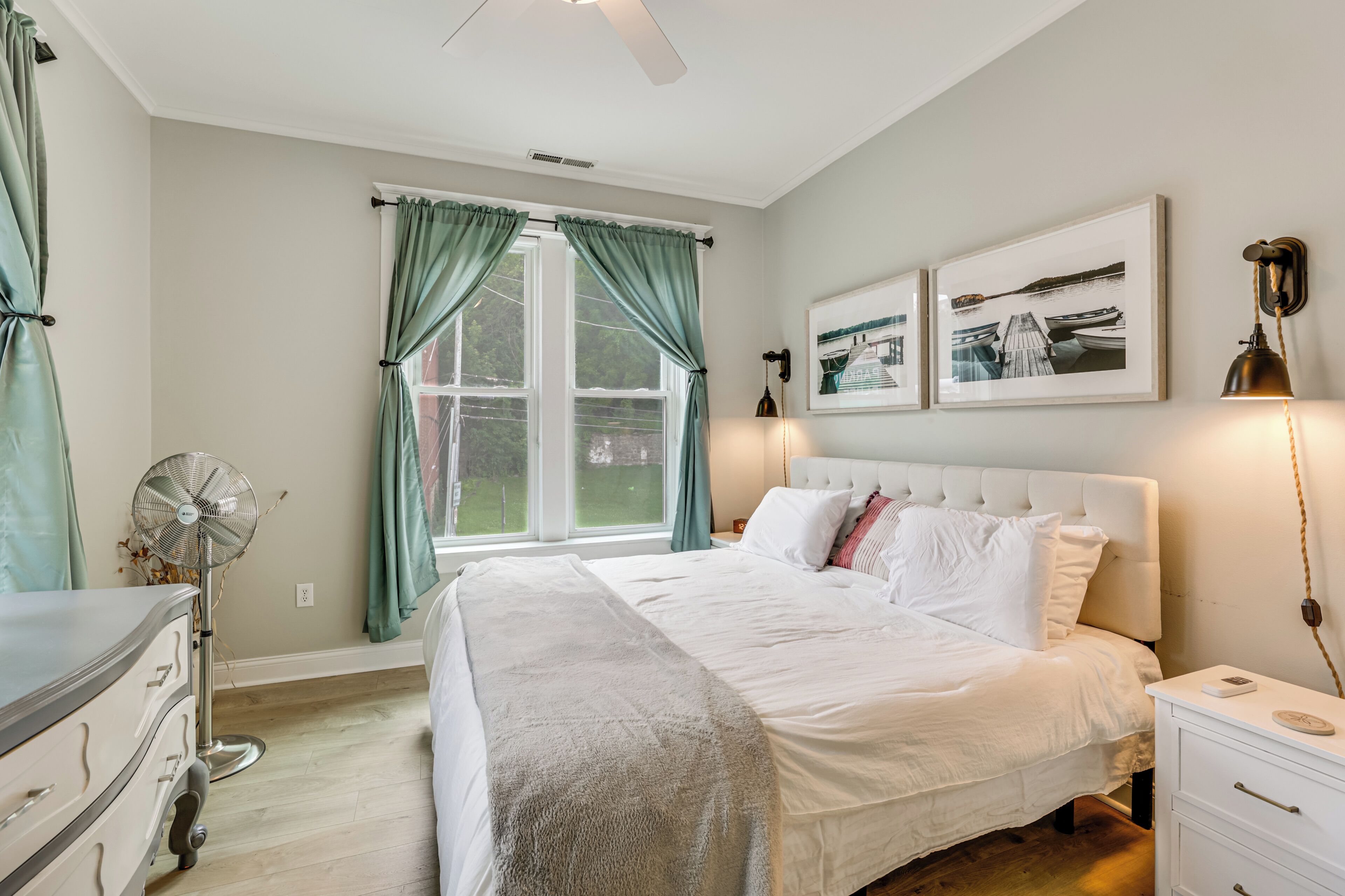 A bedroom with a bed, a dresser, a nightstand, a fan, and a window with curtains.