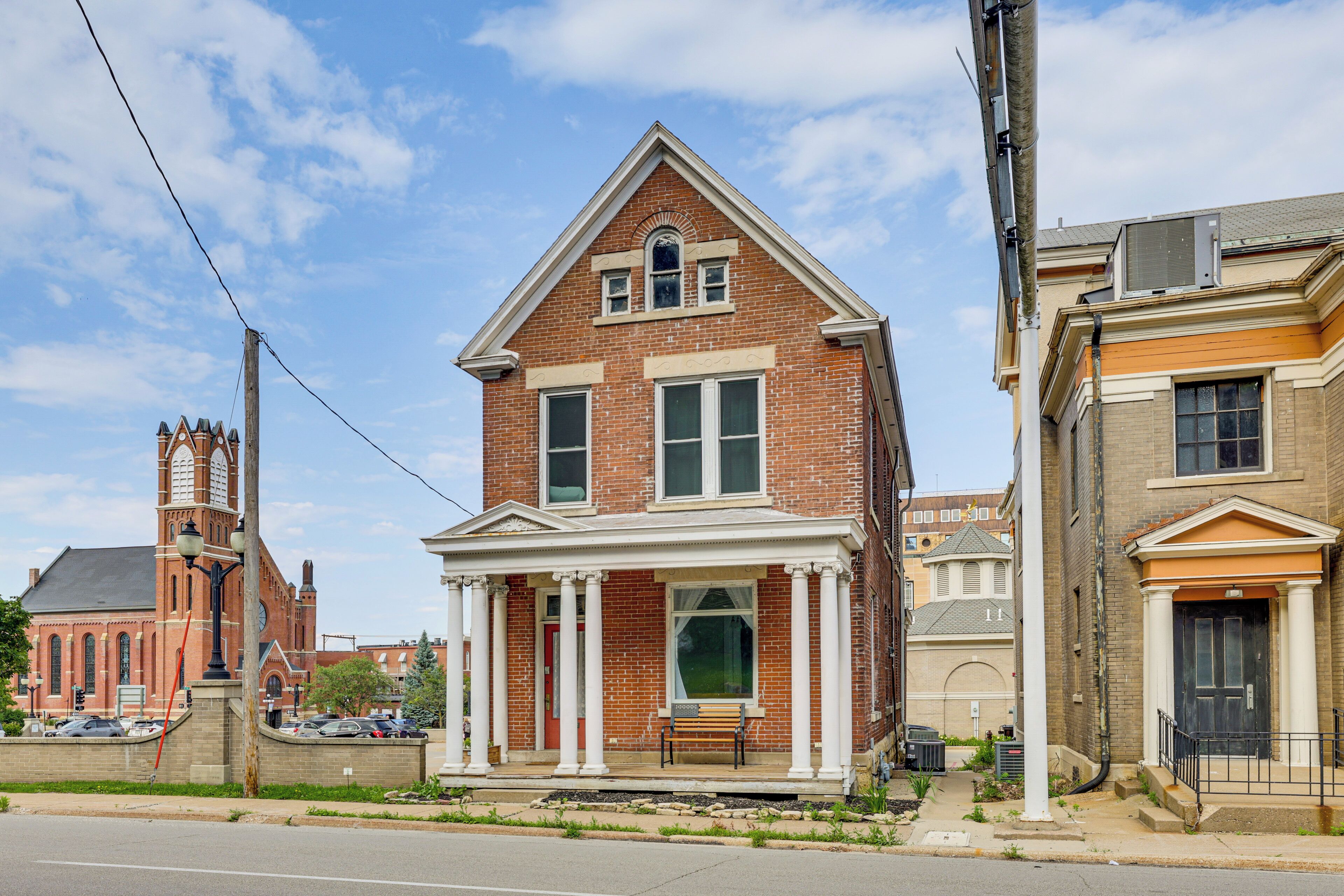 A two-story brick house with a front porch, a red door, and a small front yard.