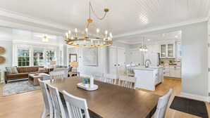A spacious living room with a large wooden dining table, white chairs, and a chandelier. - Seabrook's Someplace Else: Hot Tub, Pet Free, Steps From Town Center! (Pacific Beach)