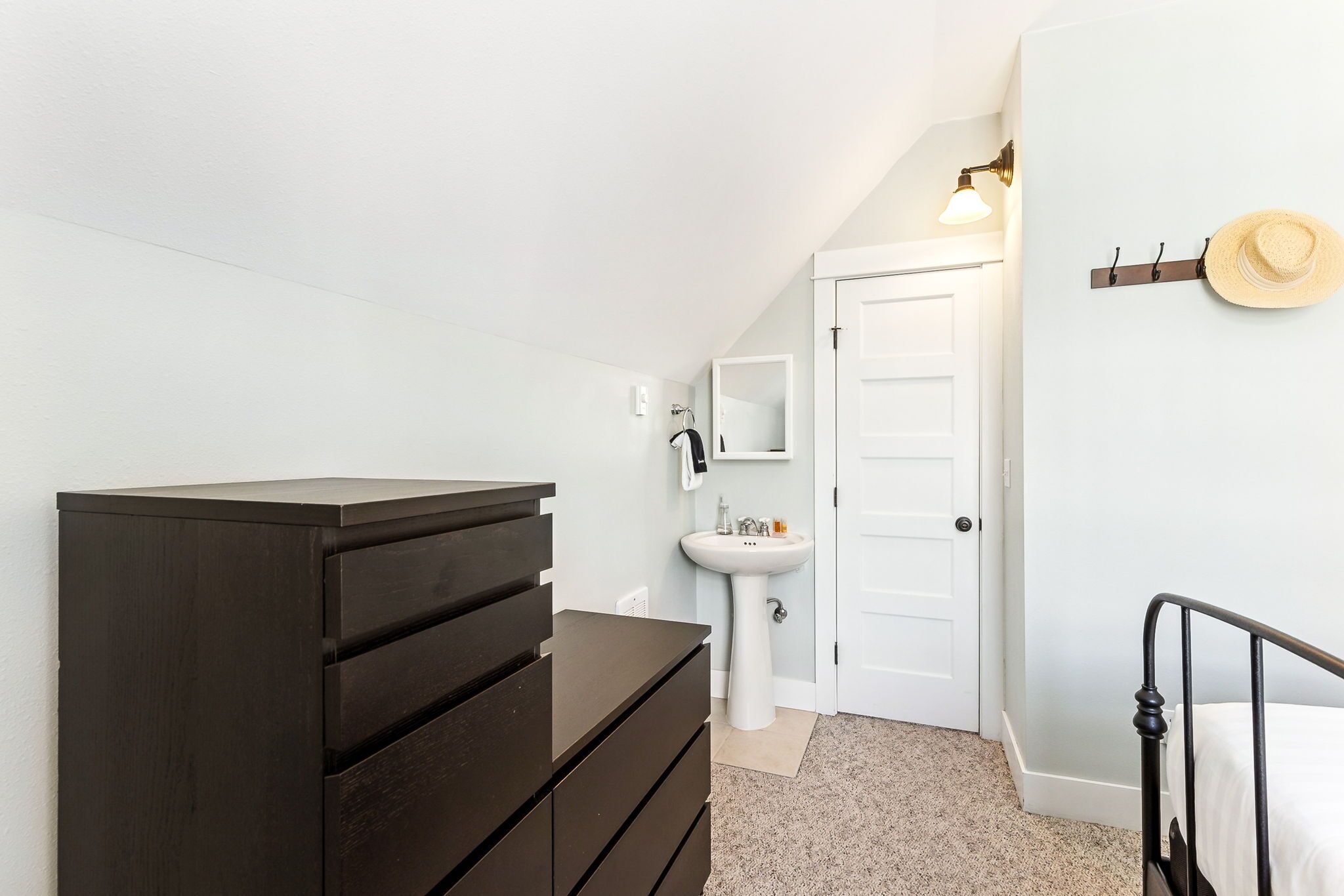 Une chambre avec une commode en bois foncé, une porte blanche, un mur blanc, un petit lavabo et un chapeau accroché au mur.