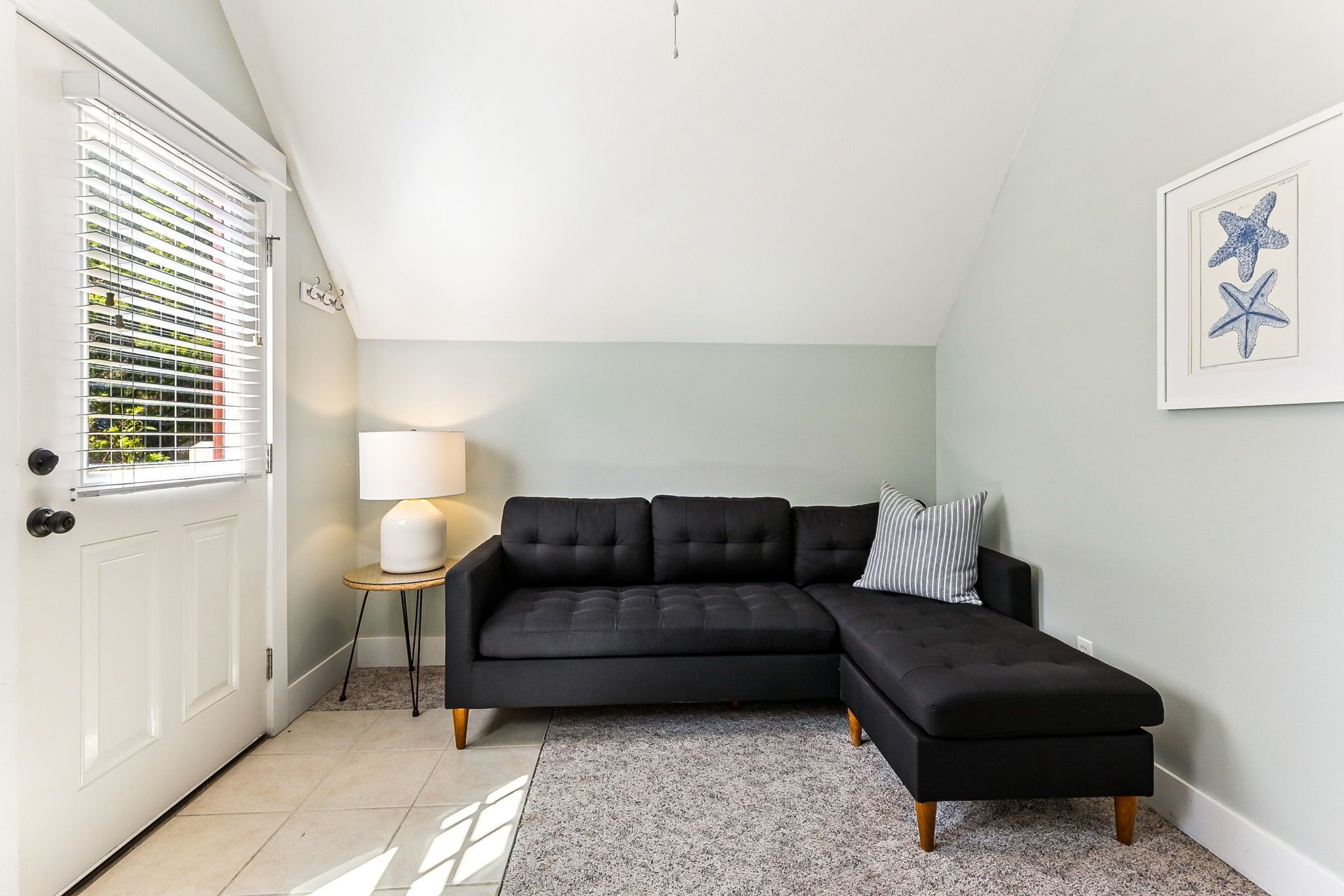 Ein modernes Wohnzimmer mit einem schwarzen modularen Sofa, einem kleinen Beistelltisch mit einer Lampe und einem Bilderrahmen mit einer Meerstern-Druckgrafik an der Wand.