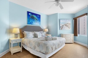 A bedroom with a bed, two bedside tables, a ceiling fan, and artwork on the walls. - Sea Turtle Retreat! 817 END UNIT w/ Wrap Around Balcony and Panoramic Views! (Panama City Beach)