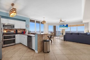 A modern kitchen with white cabinets, a stainless steel oven, and a large island. There is a dining area with a blue sofa and a view of the ocean through large windows. - Sea Turtle Retreat! 817 END UNIT w/ Wrap Around Balcony and Panoramic Views! (Panama City Beach)