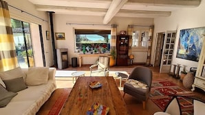 A spacious living room with a wooden coffee table, a sofa, and a chair. There is a fireplace, a bookshelf, and a large window with a view of the outdoors. - La Villa de Robion in Robion (Robion)