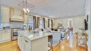 A modern kitchen with a central island, stainless steel appliances, and a dining area with a sofa and table. - Cozy 5BR Jamaica Plain | Walk to T & Downtown (Boston)
