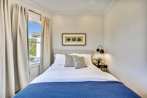 A neatly made bed with a blue bedspread, a framed picture on the wall, and a nightstand with a lamp and a small plant. - Cozy 5BR Jamaica Plain | Walk to T & Downtown (Boston)