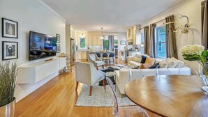 A modern living room with a TV, sofa, dining table, and kitchen in the background. - Cozy 5BR Jamaica Plain | Walk to T & Downtown (Boston)