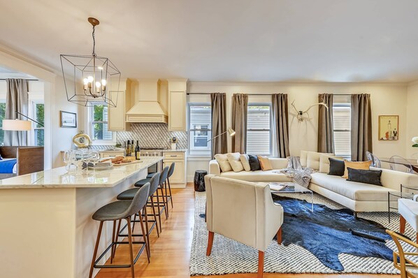 A modern kitchen with a central island, a dining area with bar stools, a living room with a sofa and a large rug, and a dining table with chairs. - Cozy 5BR Jamaica Plain | Walk to T & Downtown (Boston)