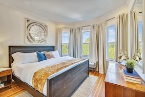 A bedroom with a bed, a nightstand, a desk, a window with curtains, and a decorative wall piece.