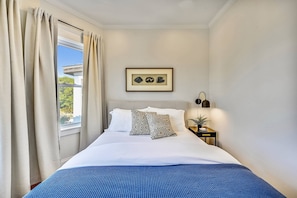 A neatly made bed with a blue bedspread, a framed picture on the wall, and a nightstand with a lamp and a small plant. - Cozy 5BR Jamaica Plain | Walk to T & Downtown (Boston)