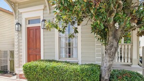 Une maison avec une porte d’entrée en bois, une fenêtre ornée de rideaux en dentelle et un arbre devant.
