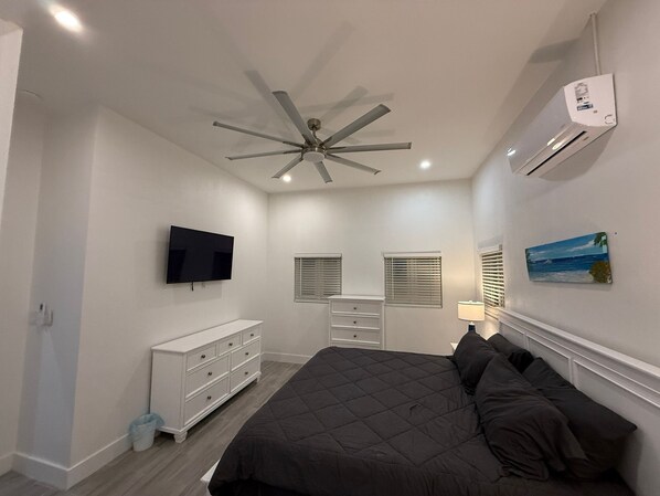 Un dormitorio con cama, ventilador de techo, aire acondicionado, televisión, cómoda y un cuadro en la pared.