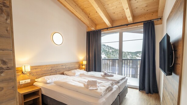 Un dormitorio con techo de madera, una cama con ropa de cama blanca, una mesita de noche con lámpara, un televisor montado en la pared y un balcón con cortinas.