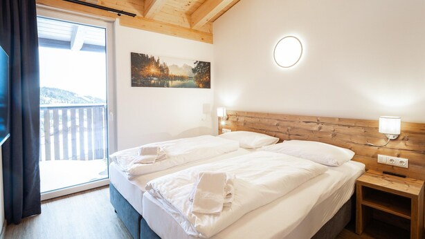 A hotel room with a wooden ceiling, a large bed with white linens, a painting of a landscape, a nightstand with a lamp, and a view of a balcony. - Spacious 4-bedroom Penthouse in Katschberghohe (Rennweg am Katschberg)