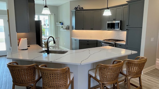 A modern kitchen with grey cabinets, a large island with bar stools, a built-in microwave, and pendant lighting. - Stunning home with water view & golf cart Winter Cancellation - Reduced Rates (The Villages)