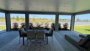 A spacious room with a large window offering a view of a golf course and a lake. There is a dining area with a table and chairs, and a seating area with a sofa. - Stunning home with water view & golf cart Winter Cancellation - Reduced Rates (The Villages)