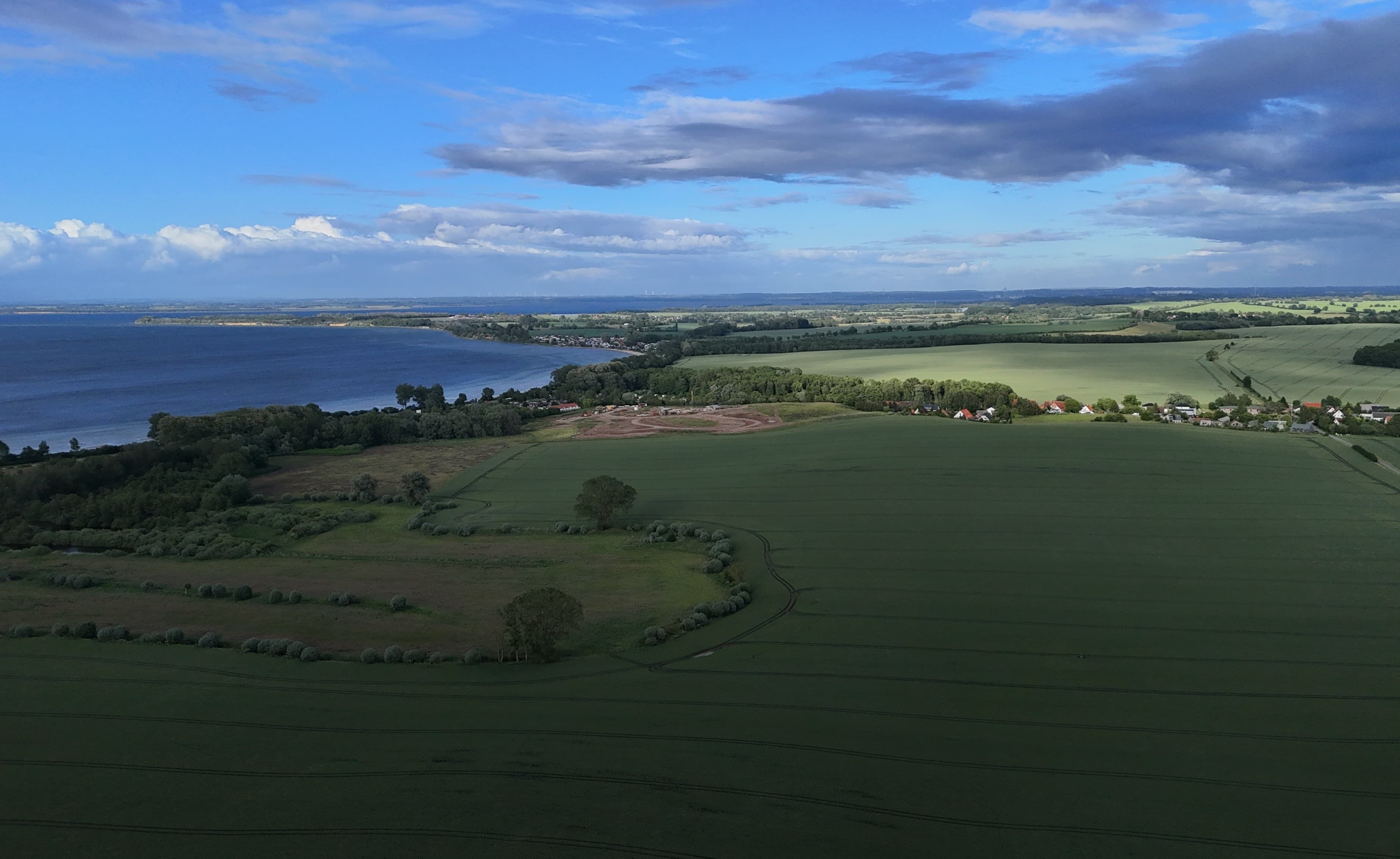 Luftaufnahme einer ländlichen Landschaft mit Feldern, einem Gewässer und verstreuten Häusern.