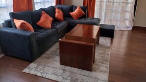 Ein Wohnzimmer mit einem dunkelgrauen Sofa, orangefarbenen Kissen, einem Holztisch und einem gemusterten Teppich.