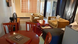 Ein kompakter Wohnbereich mit Esstisch, zwei Sofas, einer kleinen Küchenzeile und einem Fenster mit Vorhängen.