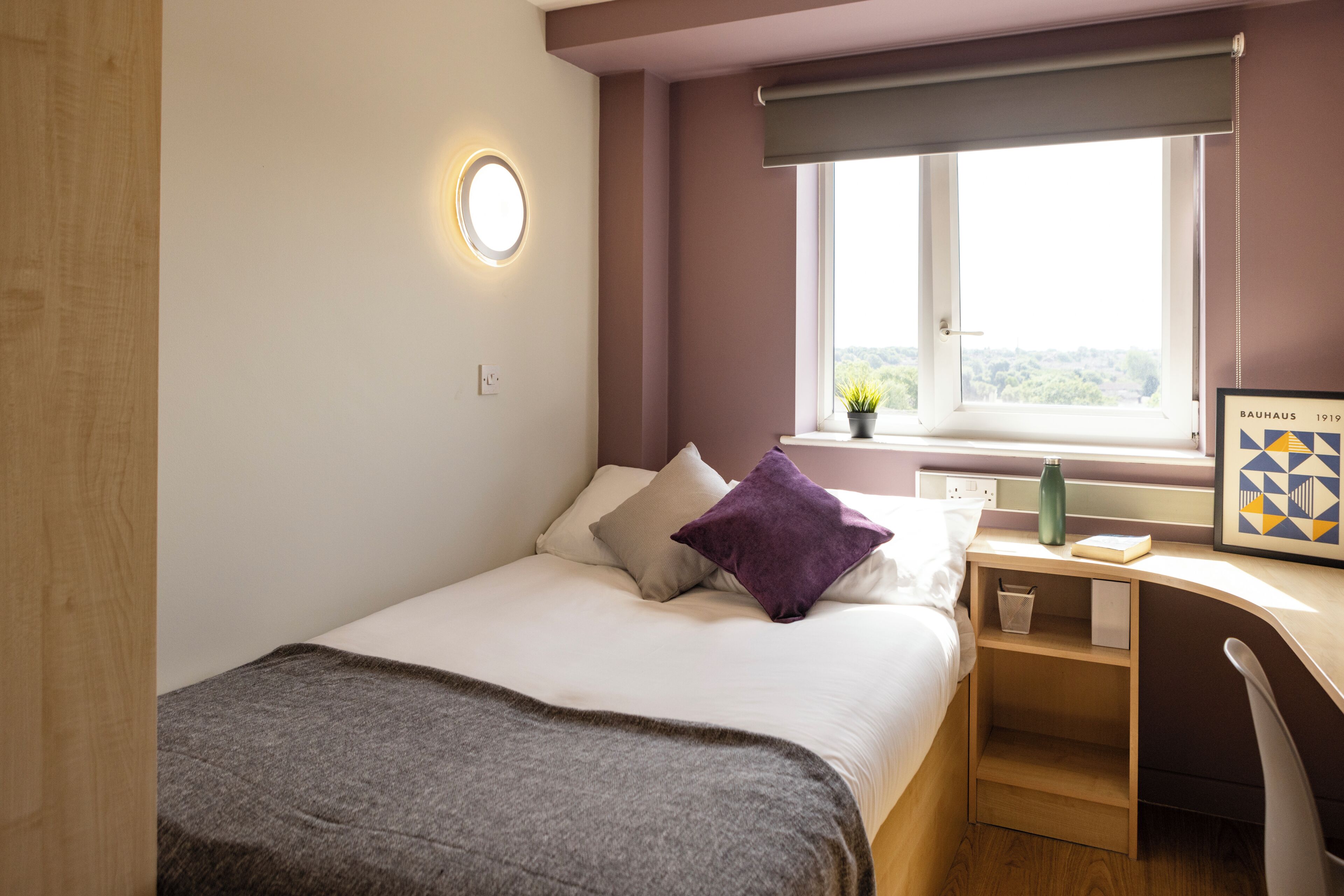 A bedroom with a bed, a desk, a window with a view, and a wall-mounted light.