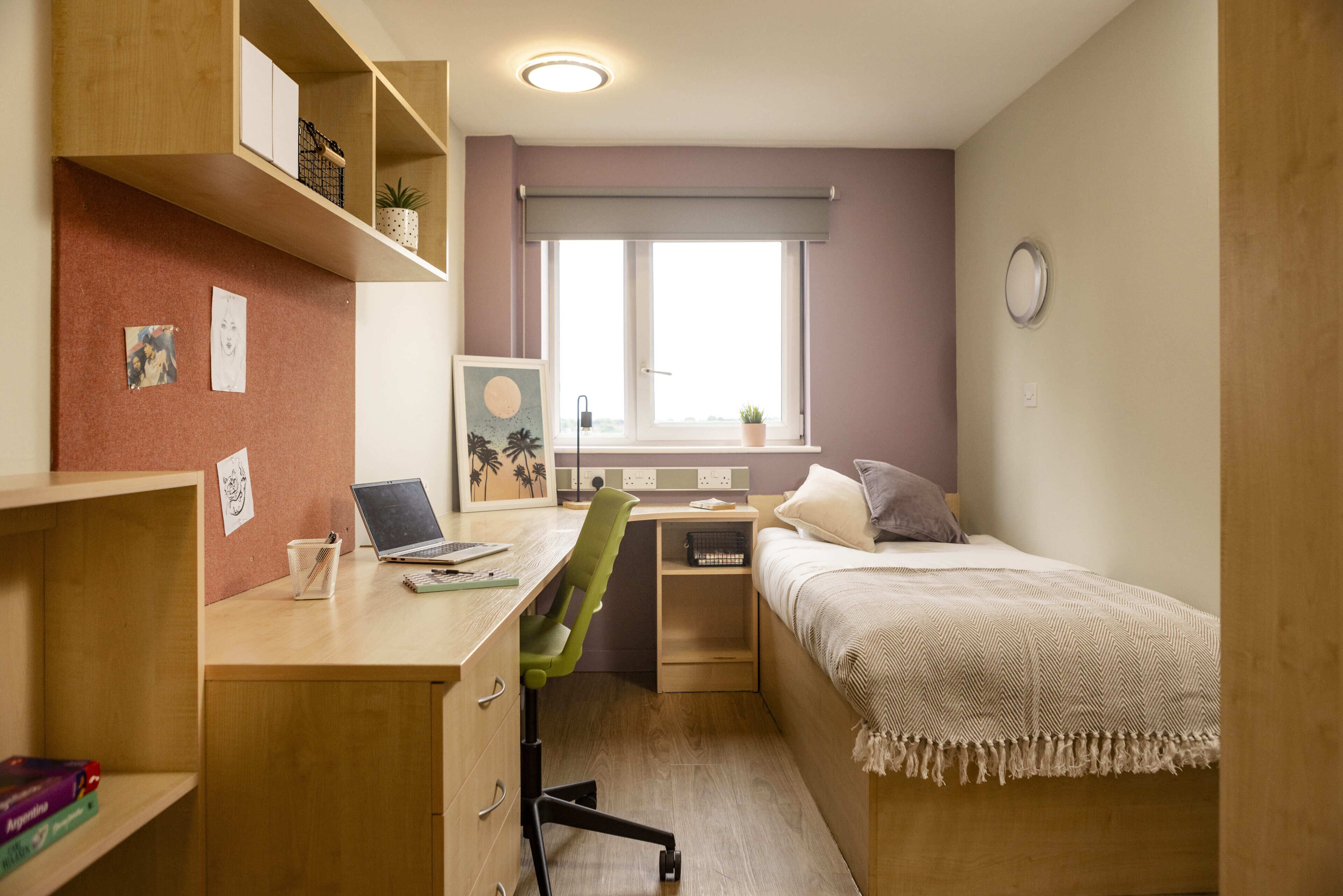 Une pièce de taille modeste, comprenant un bureau, un lit, une fenêtre donnant sur un paysage agréable et une petite étagère avec quelques objets.