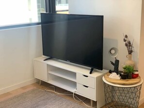 Un televisor de pantalla plana sobre un mueble blanco, una mesita redonda con una lámpara y adornos, y una ventana con vistas.