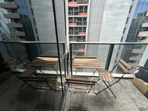 A balcony with wooden tables and chairs, a glass railing, and a view of a building with balconies. - Beautiful and Convenient apartment in CBD (Melbourne)
