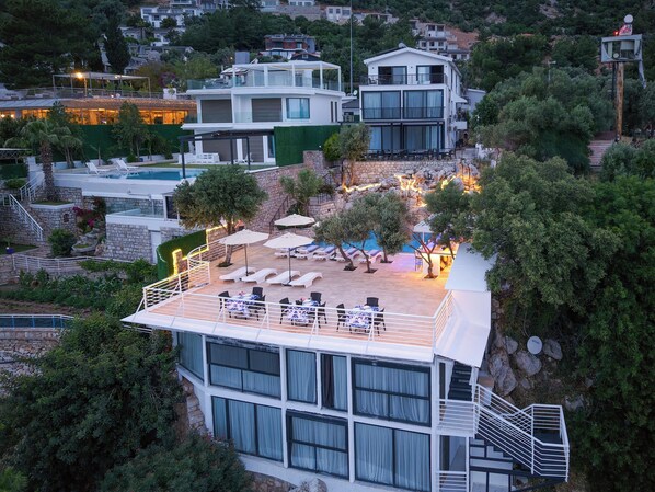 Un edificio moderno a più piani con area piscina sul tetto, comprensiva di lettini e ombrelloni, circondata da vegetazione lussureggiante e edifici residenziali sullo sfondo.