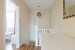 En gang med et hvidt gelænder, en dør der fører til et værelse med en seng, og en lampe på væggen.