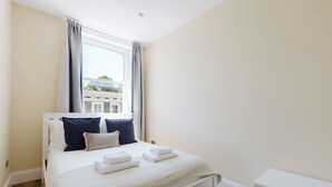 Ein Schlafzimmer mit einem Bett, weißer Bettwäsche, einem Fenster mit Vorhängen und Blick auf Gebäude.
