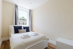 A bedroom with a bed, white bedding, a window with curtains, and a view of buildings outside. - Newly Renovated 2BD Top Floor Flat, West Brompton (Greater London)