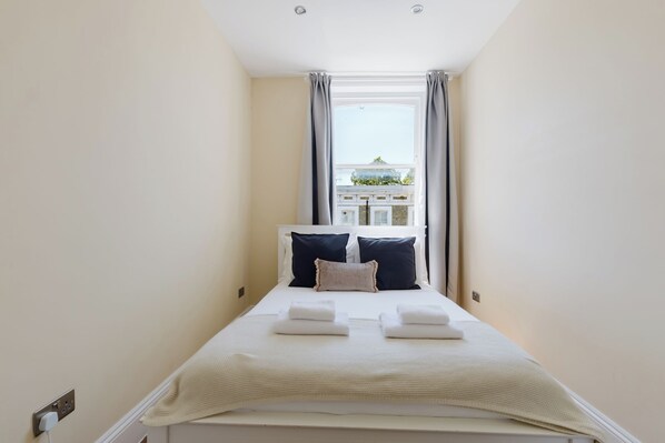 A narrow hotel room with a bed, two pillows, and a window with curtains.