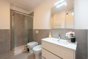 A modern bathroom with a glass-enclosed shower, a toilet, a bidet, a white sink with a mirror, and a vase with flowers. - Central Apartment Piazza Dante Genova (Genoa)