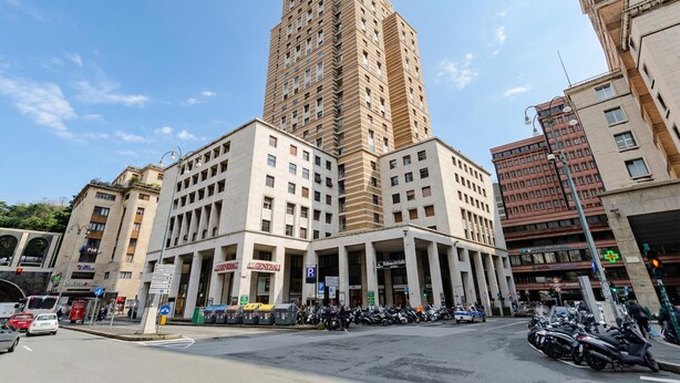 A multi-story building with a prominent clock tower, a parking area with motorcycles, and various commercial establishments. - Central Apartment Piazza Dante Genova (Genoa)