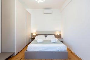 Quarto de hotel moderno com cama, mesas de cabeceira, um banco e um aparelho de ar condicionado.