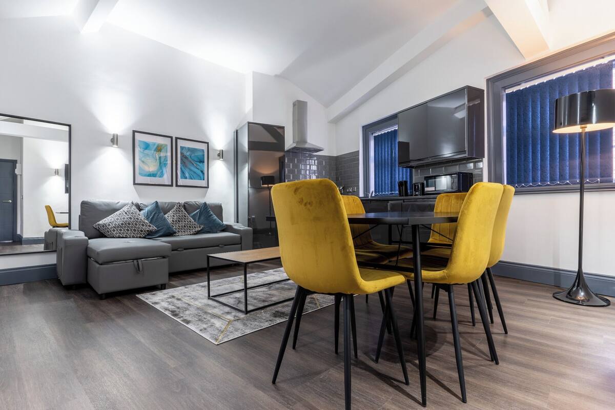 A modern living room with a gray sofa, yellow chairs, a dining table, and a kitchen area with a TV and appliances.