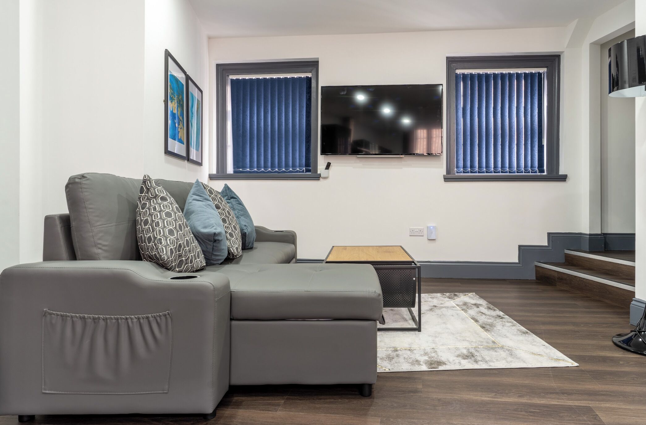 Una sala moderna con un sofá seccional gris, dos ventanas con cortinas azules, un televisor de pantalla plana y una pequeña mesa de madera.
