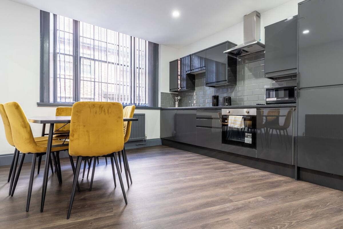 A modern kitchen with grey cabinets, a built-in oven, and a dining area with yellow chairs.