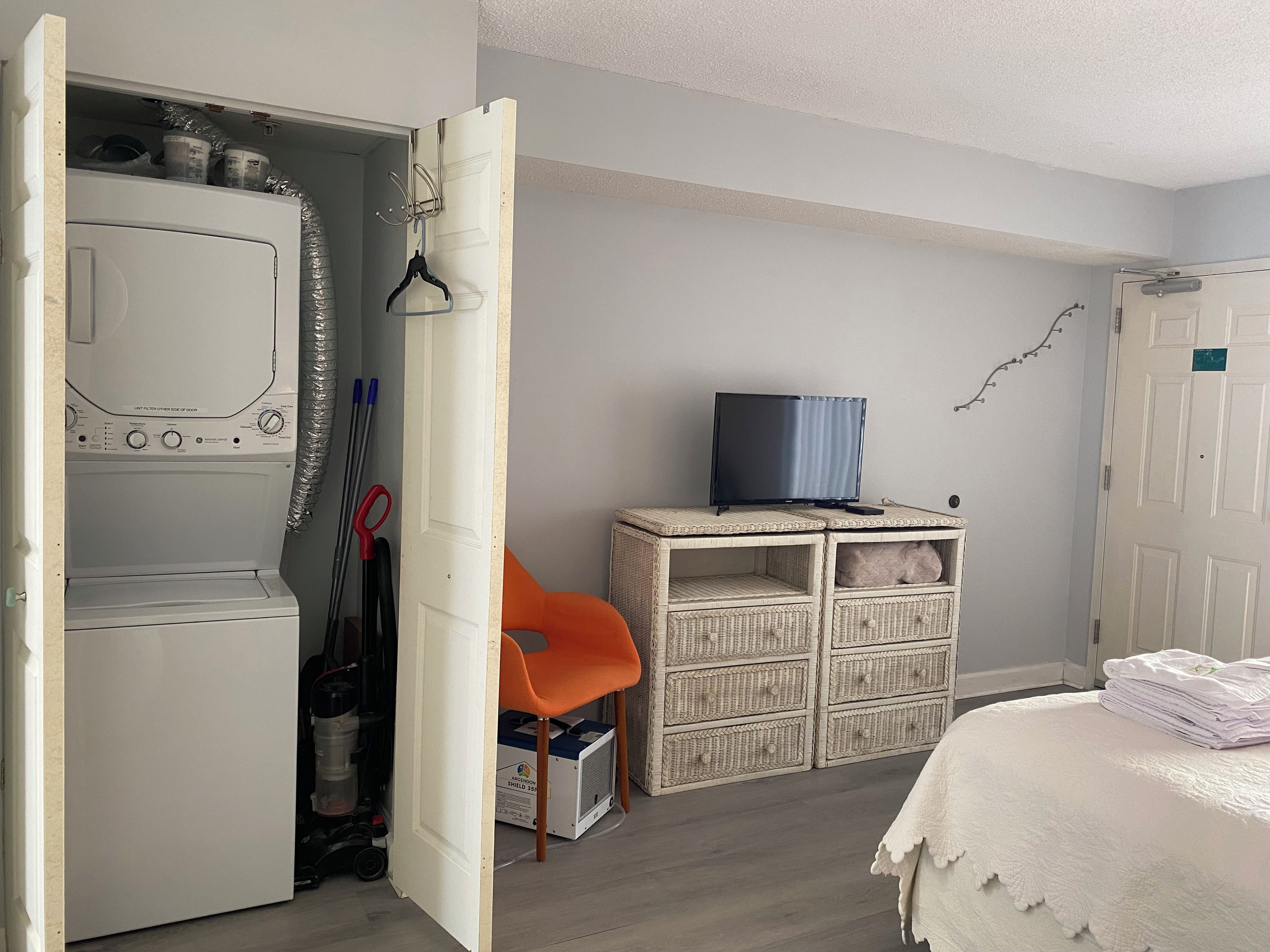 Una habitación con lavadora, televisor sobre un mueble bajo, muebles de mimbre y una cama con cubrecama blanco.