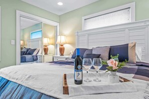 A neatly made bed with a bottle of wine and glasses, a mirror, and a window with blinds.