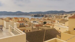 Un paysage urbain côtier avec des bâtiments aux toits de tuiles rouges, une vue dégagée sur la mer et des montagnes lointaines.
