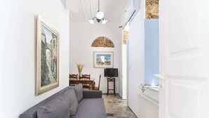 Un couloir étroit avec un canapé gris, une table à manger avec des chaises, un téléviseur posé sur un meuble et une porte donnant sur une autre pièce.