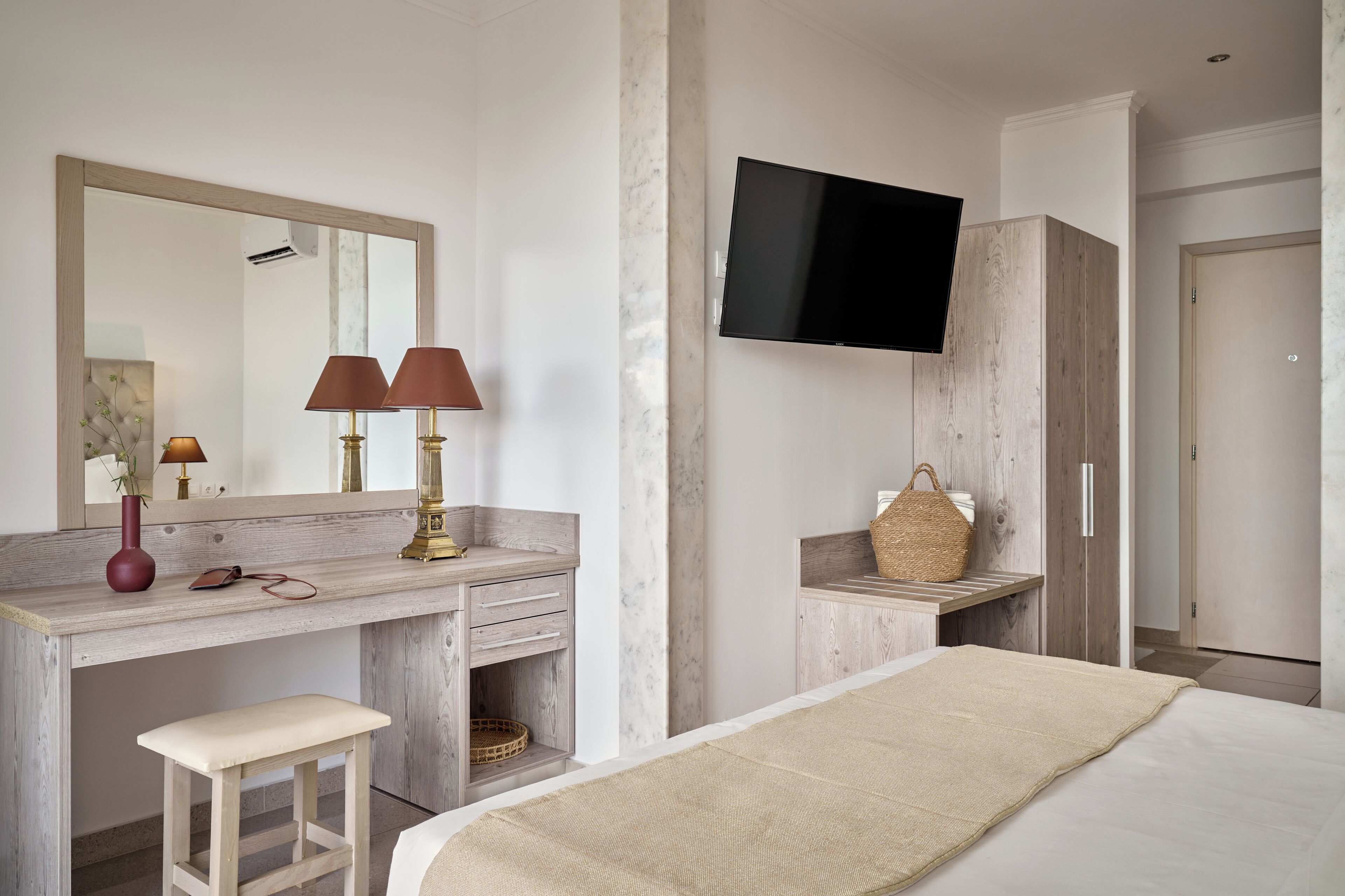 A hotel room with a bed, a desk with a mirror, a television mounted on the wall, and a wardrobe.
