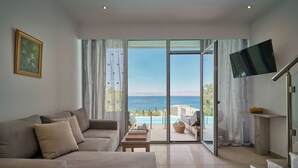 A living room with a sofa, a flat-screen TV, and a view of the sea through large glass doors. - Mythos Panorama (Corfu)