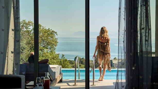 A poolside area with a view of the sea, a person walking, and a table with glasses and a bottle. - Mythos Panorama (Corfu)